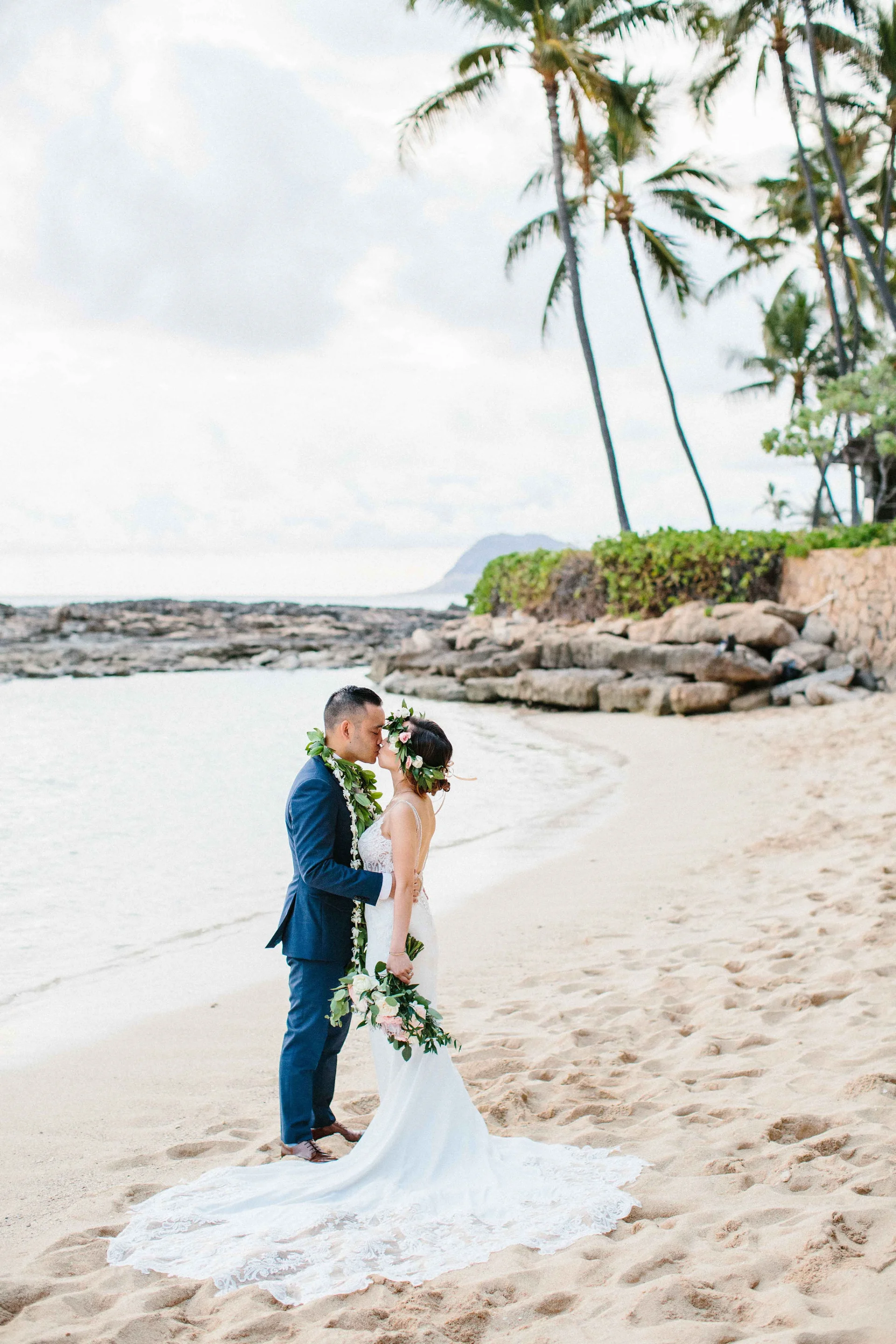 Bride and groom embracing on Lanikūhonua beach with lush greenery, lei, and floral crown in Oʻahu wedding setting