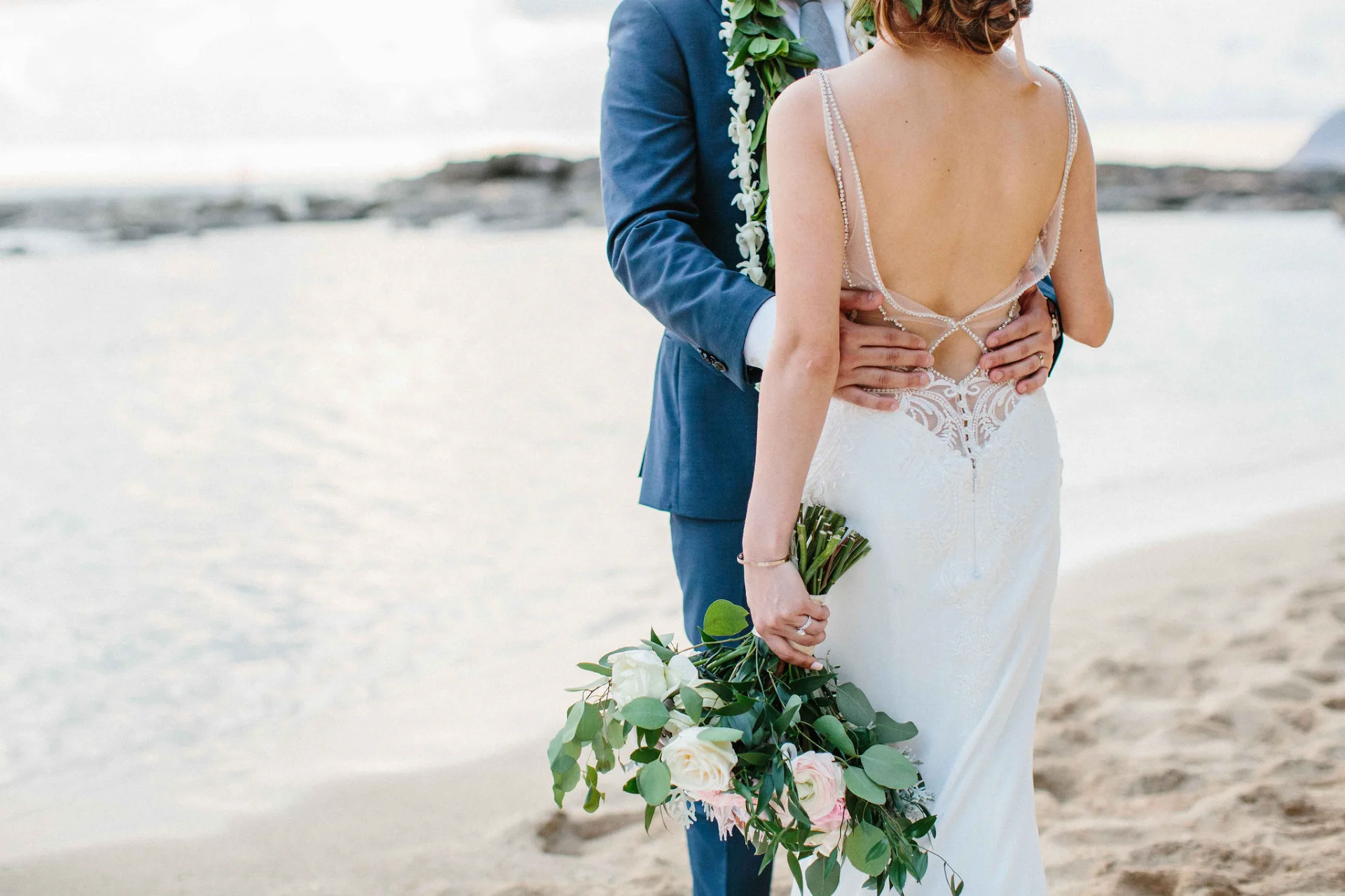 Bride in elegant lace dress holding bouquet of white and pink roses by the beach at Lanikūhonua, Oʻahu wedding.