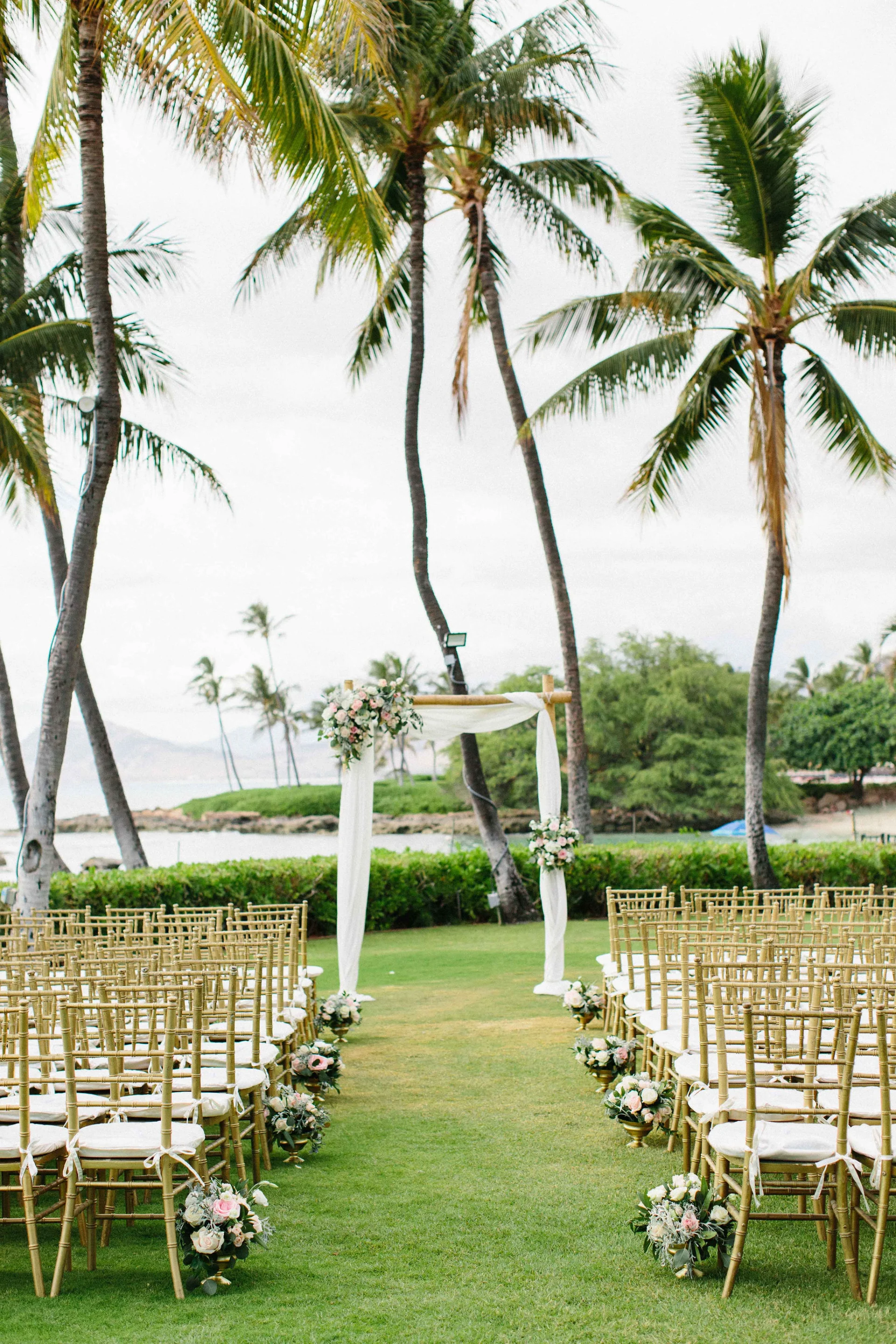 Elegant wedding ceremony setup at Lanikūhonua Cultural Institute with white floral arrangements, gold chairs, and palm trees in Oʻahu, Hawaii