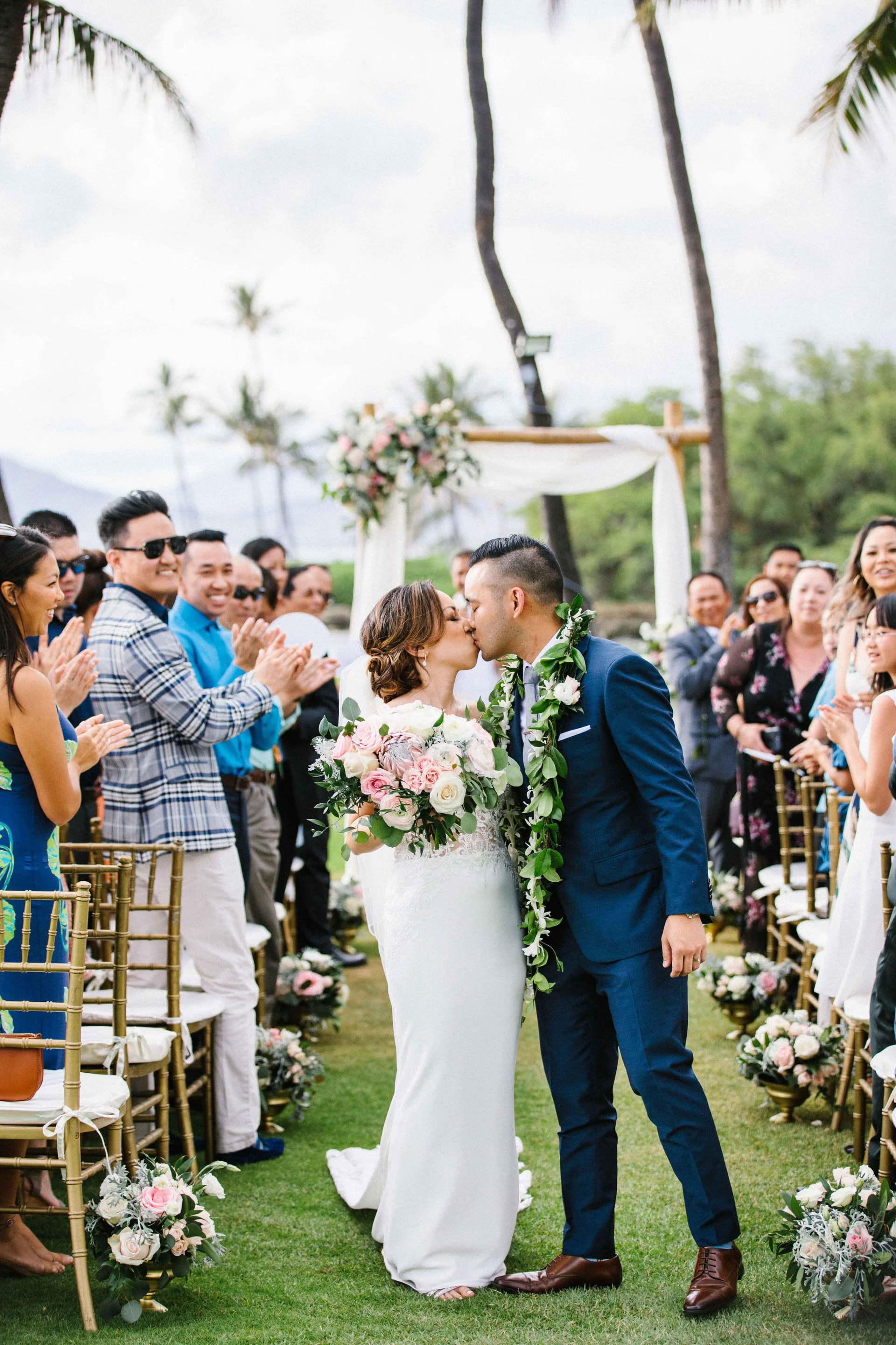 Bride and groom kissing at outdoor ceremony with pink and white rose bouquet, surrounded by guests and floral decor at Lanikūhonua, Oʻahu