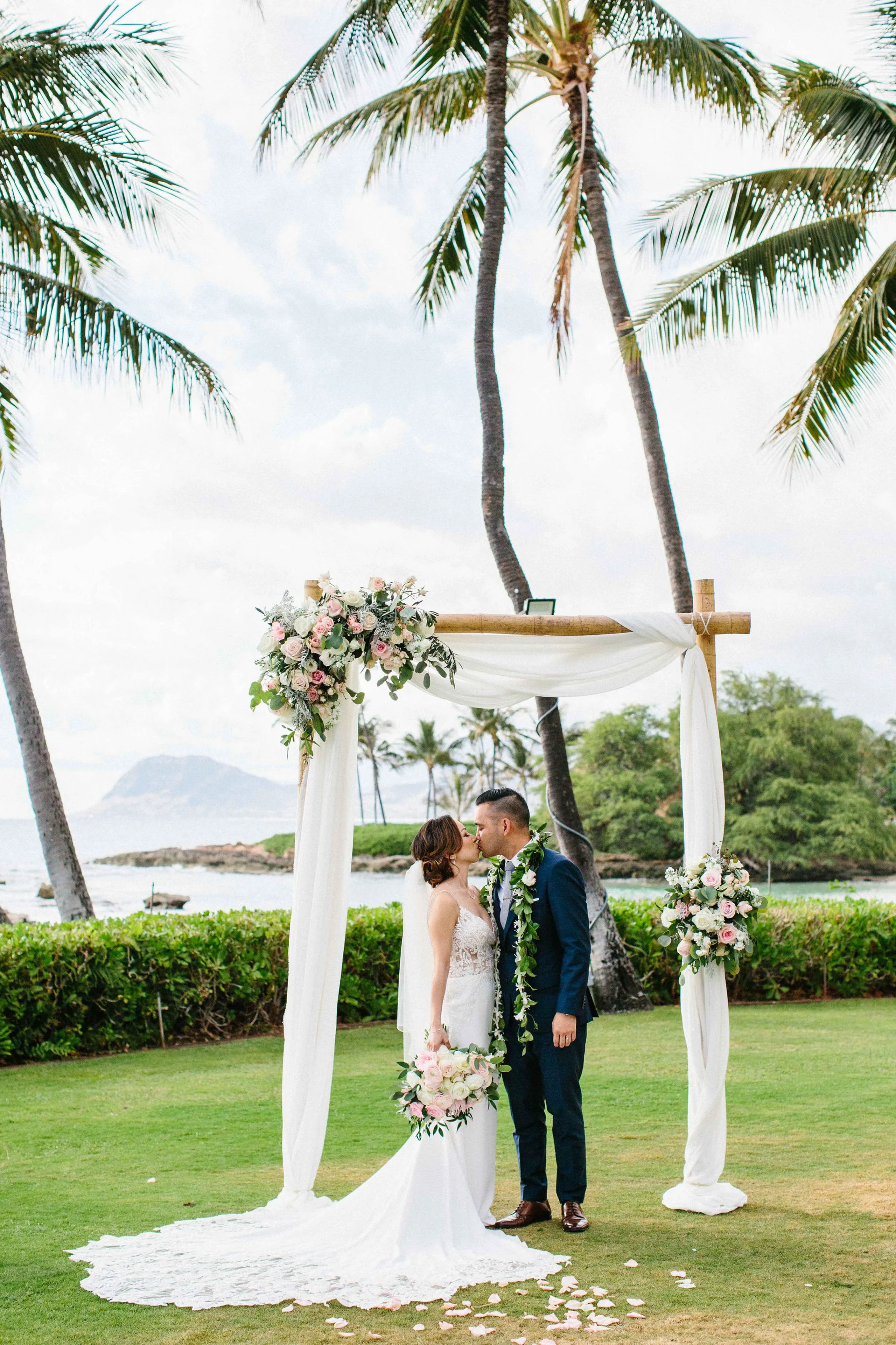 Couple kissing under a floral archway with pink roses and greenery at Lanikūhonua Cultural Institute, Oʻahu.
