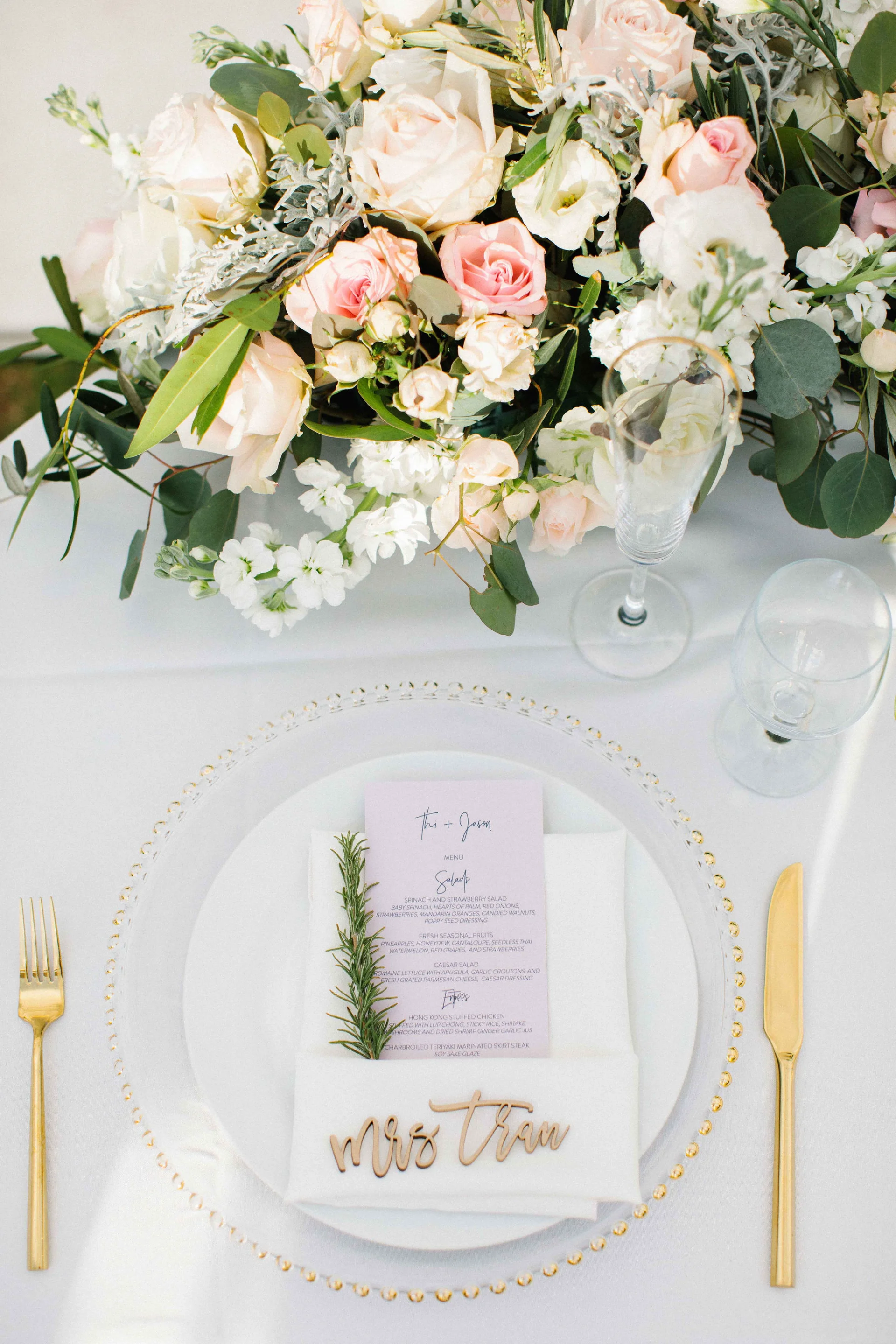 Elegant wedding table setting at Lanikūhonua with gold cutlery, floral centerpiece of pink and white roses, and personalized menu.