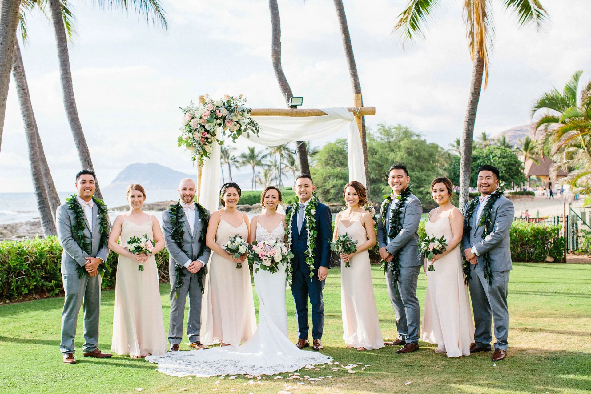 Wedding party poses under floral-decorated bamboo arch at Lanikūhonua Cultural Institute, Oʻahu, with pink and white blooms